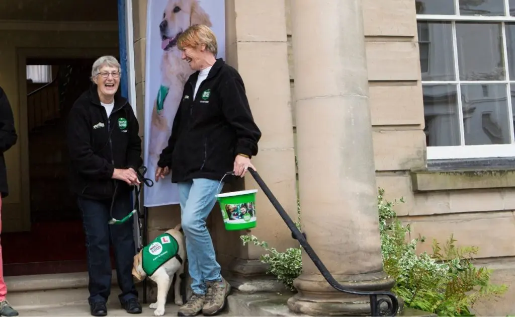 Two members of the Warwickshire supporter group outside an event with a fundraising bucket and an assistance dog