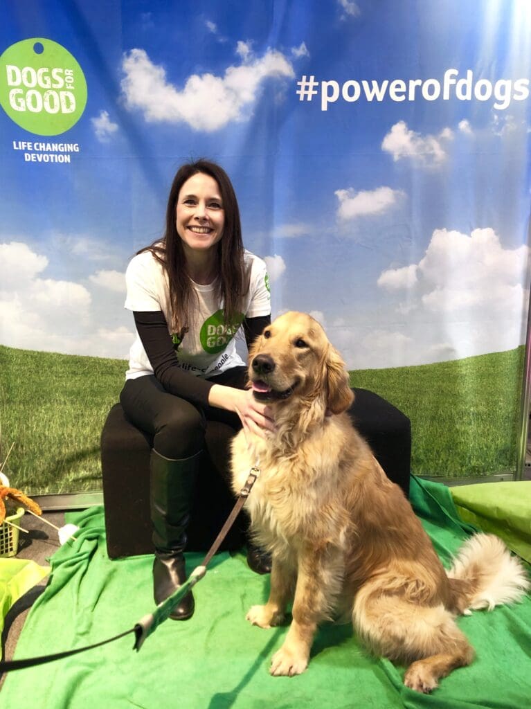 A brunette woman wearing a Dogs for Good T-shirt sits alongside a golden retriever, whose mouth is slightly open as if smiling