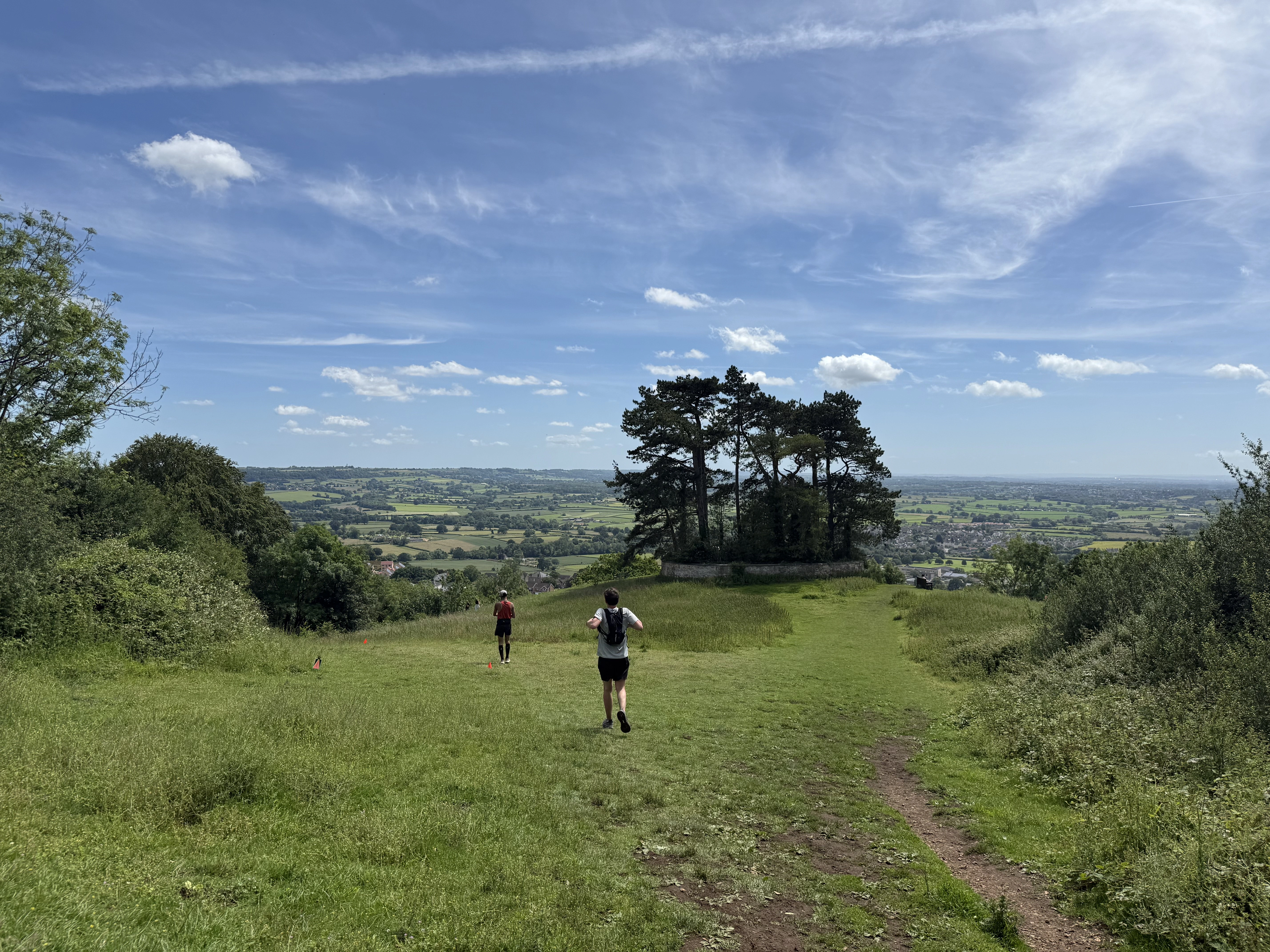 A panoramic view of the Cotswolds, with greenery, trees, fields and footpaths in the foreground and a blue sky, fields and villages in the background. Two people are running through a marked route as part of the Cotswold Way Ultra Challenge.
