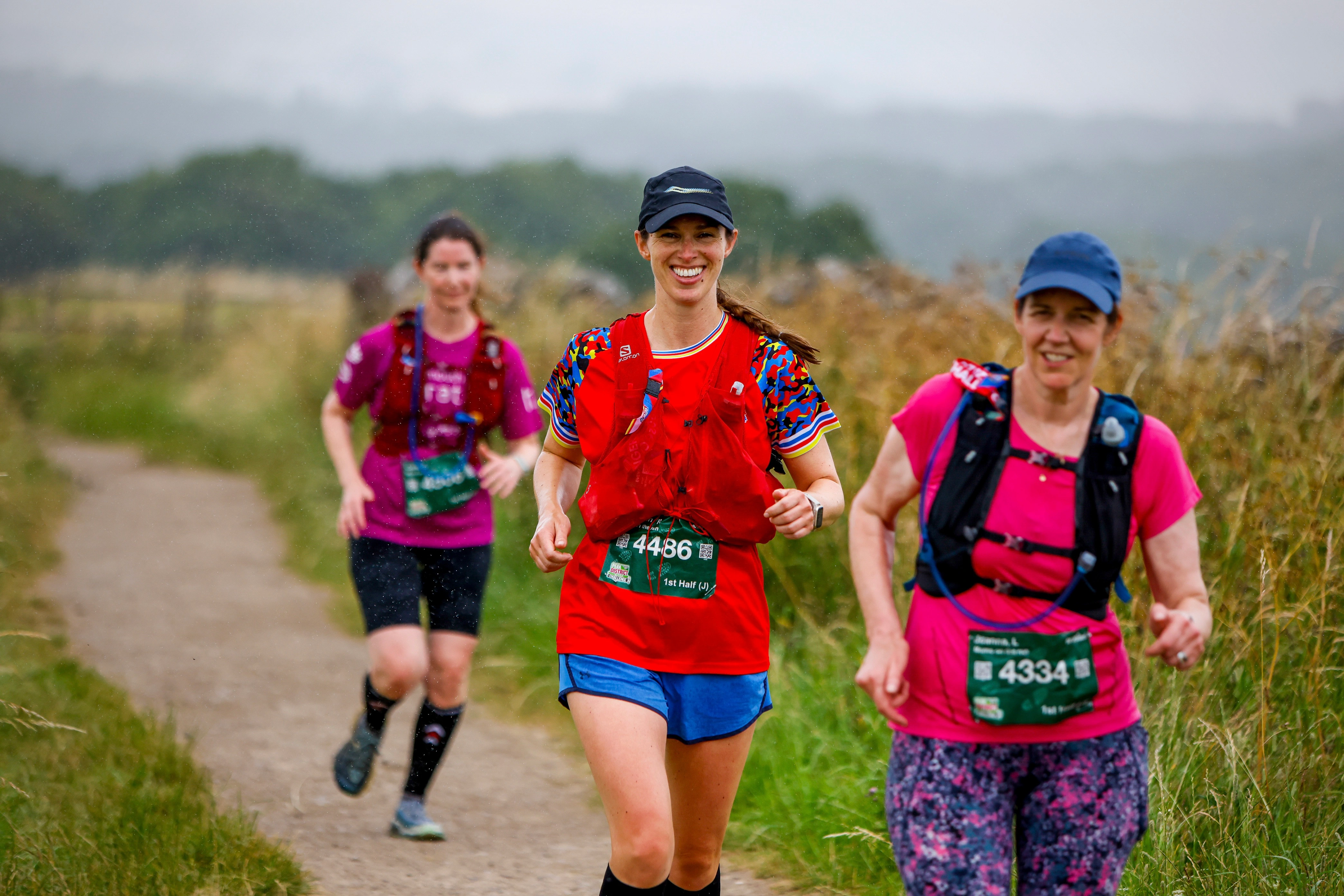 Chiltern 50 Ultra Challenge participants running along a marked path. Three women are wearing running clothes, hats and rucksacks. A view of the Chilterns can be seen in the background.