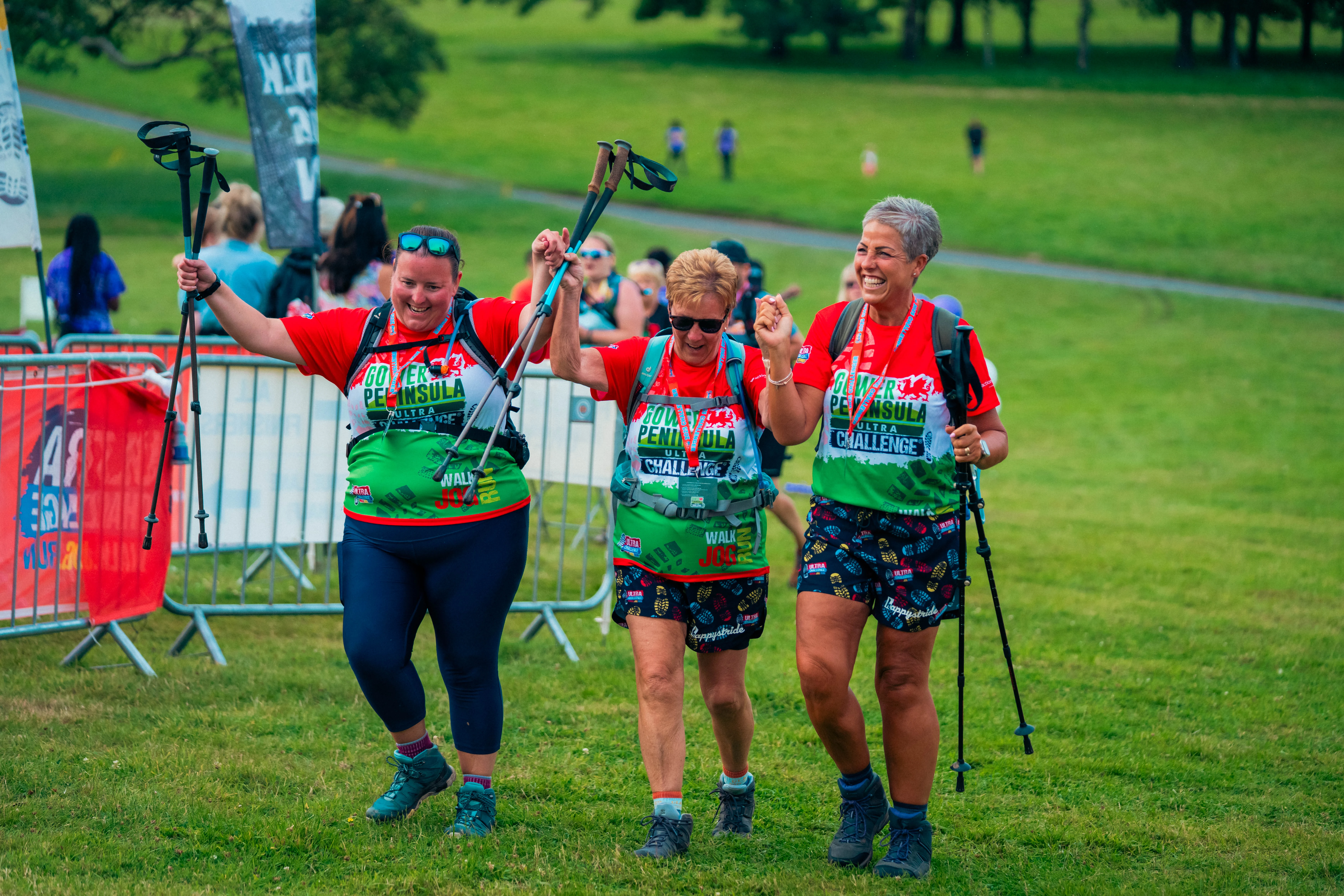 Three women celebrating as they reach the finish line of the Gower Peninsula Ultra Challenge. They are raising their walking poles in the air, holding hands and smiling. Event stewards can be seen in the background.