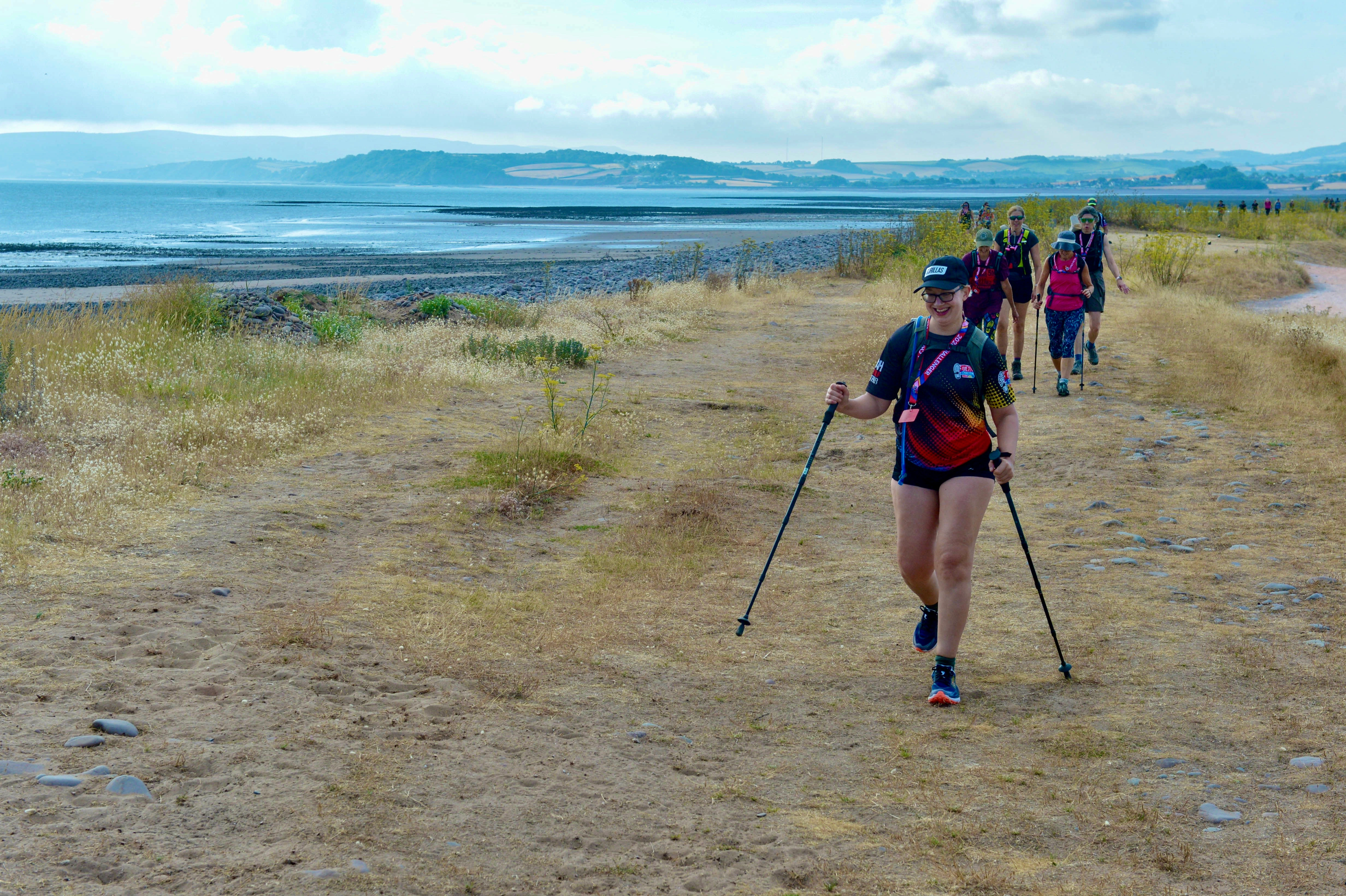 People walking along the beach during the South West Coast 50 Ultra Challenge. One person in front is smiling and using walking poles in the distance you can see other groups of walkers, the sea and the coastline of south England