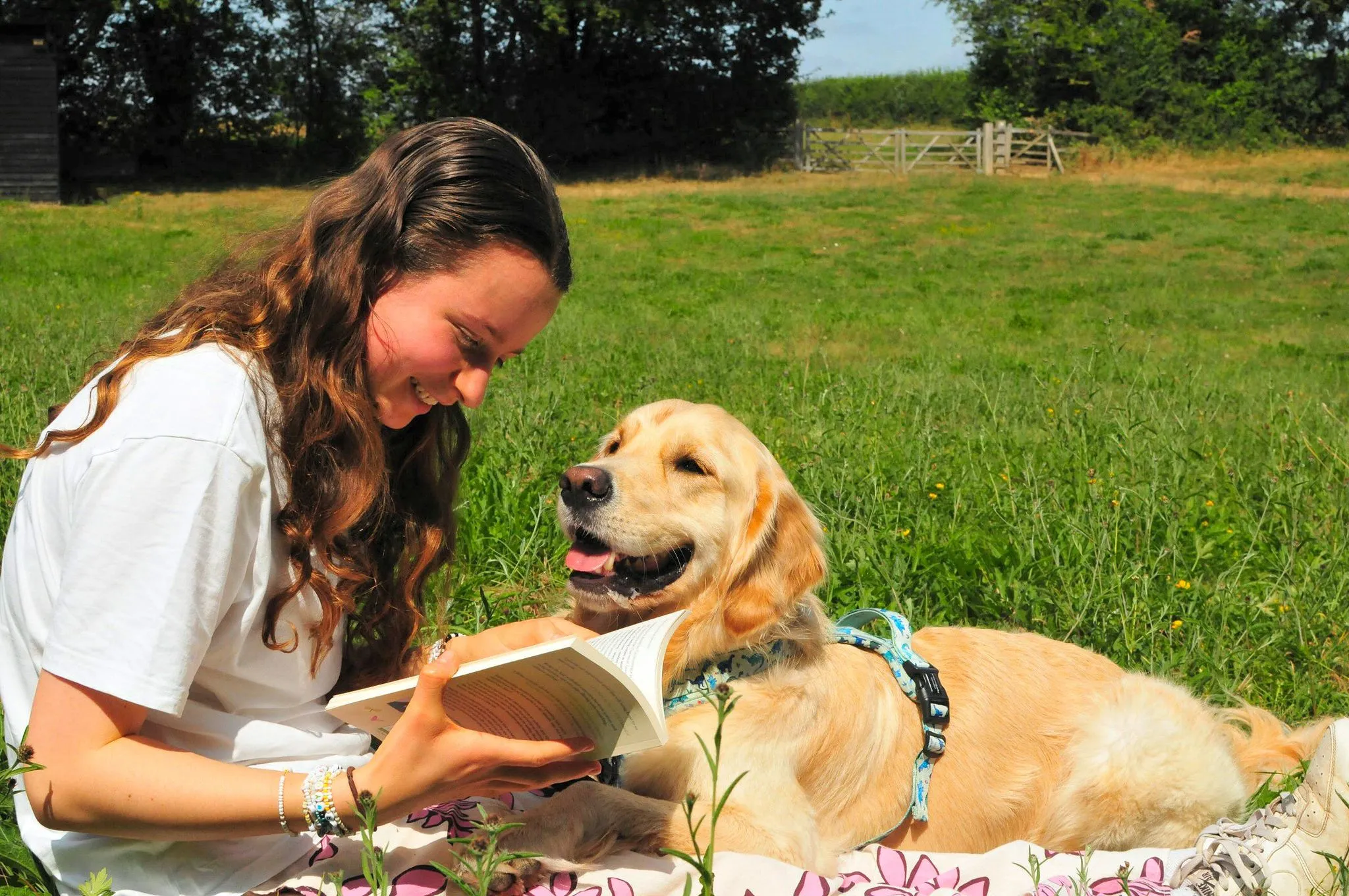 Rachel and Cruz sitting in the grass outside on a sunny day. Rachel is reading a book and Cruz is looking at her. They are sitting on a pink flowery blanket.