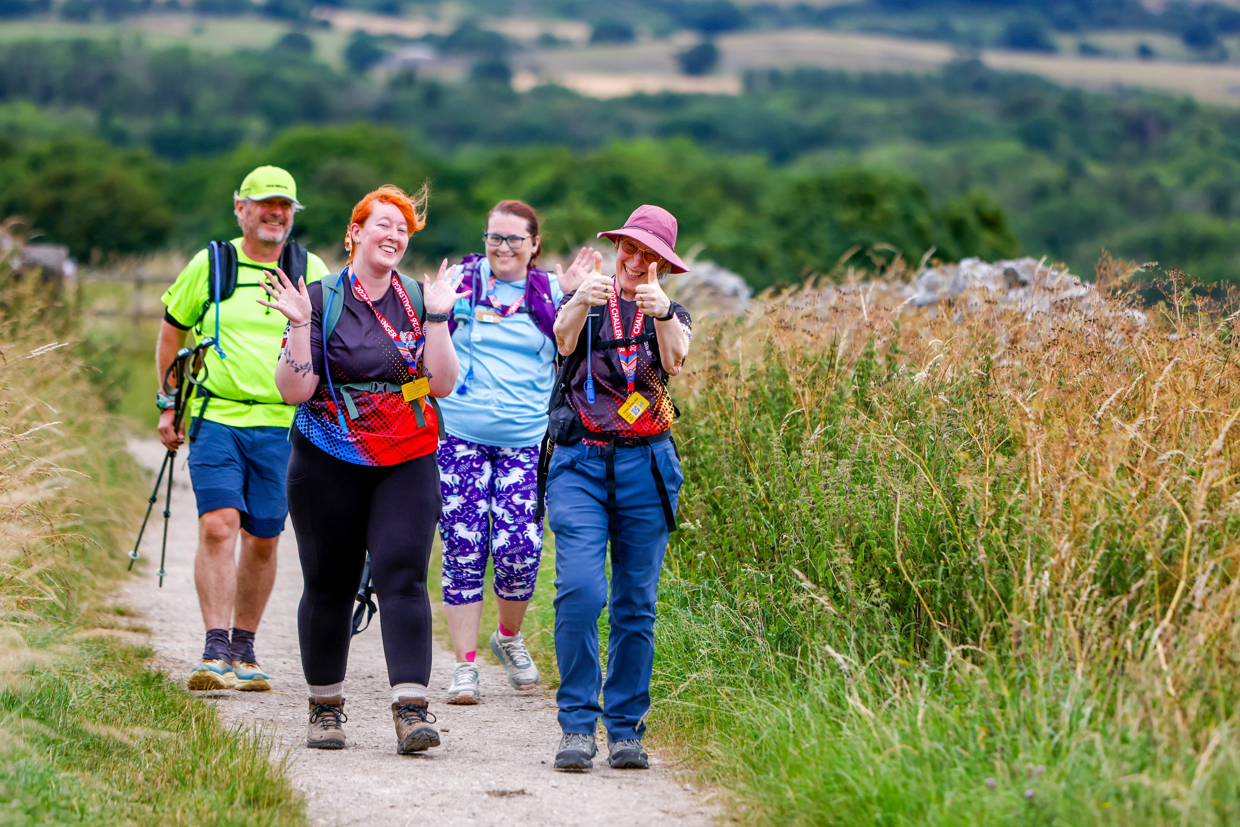 Ultra Challenge participants walking in the countryside. A group of four walkers wearing lanyards, hats and walking gear smiling and waving. They are on a country trail and fields and trees can be seen in the background.