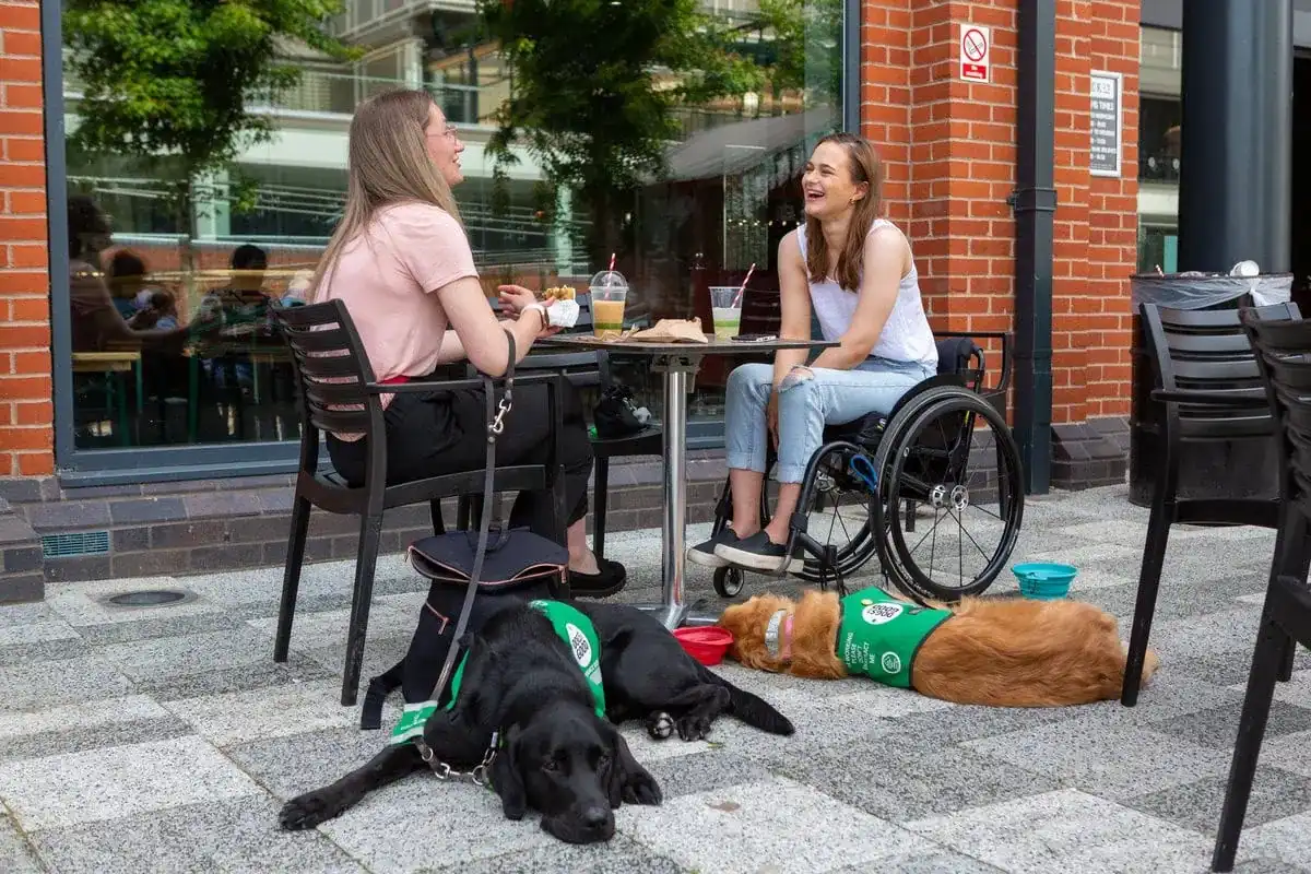 Two women sitting outside of cafe laughing and smiling while their assistance dogs lay on the ground beside them
