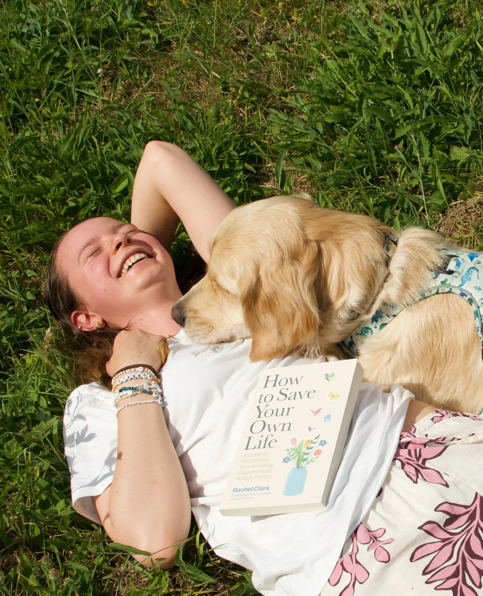 Rachel and Cruz lying down on the grass outside. Rachel is on her back with her hand behind her head and smiling, Cruz is resting on her stomach with his head on her chest. The book Rachel has written is lying next to them
