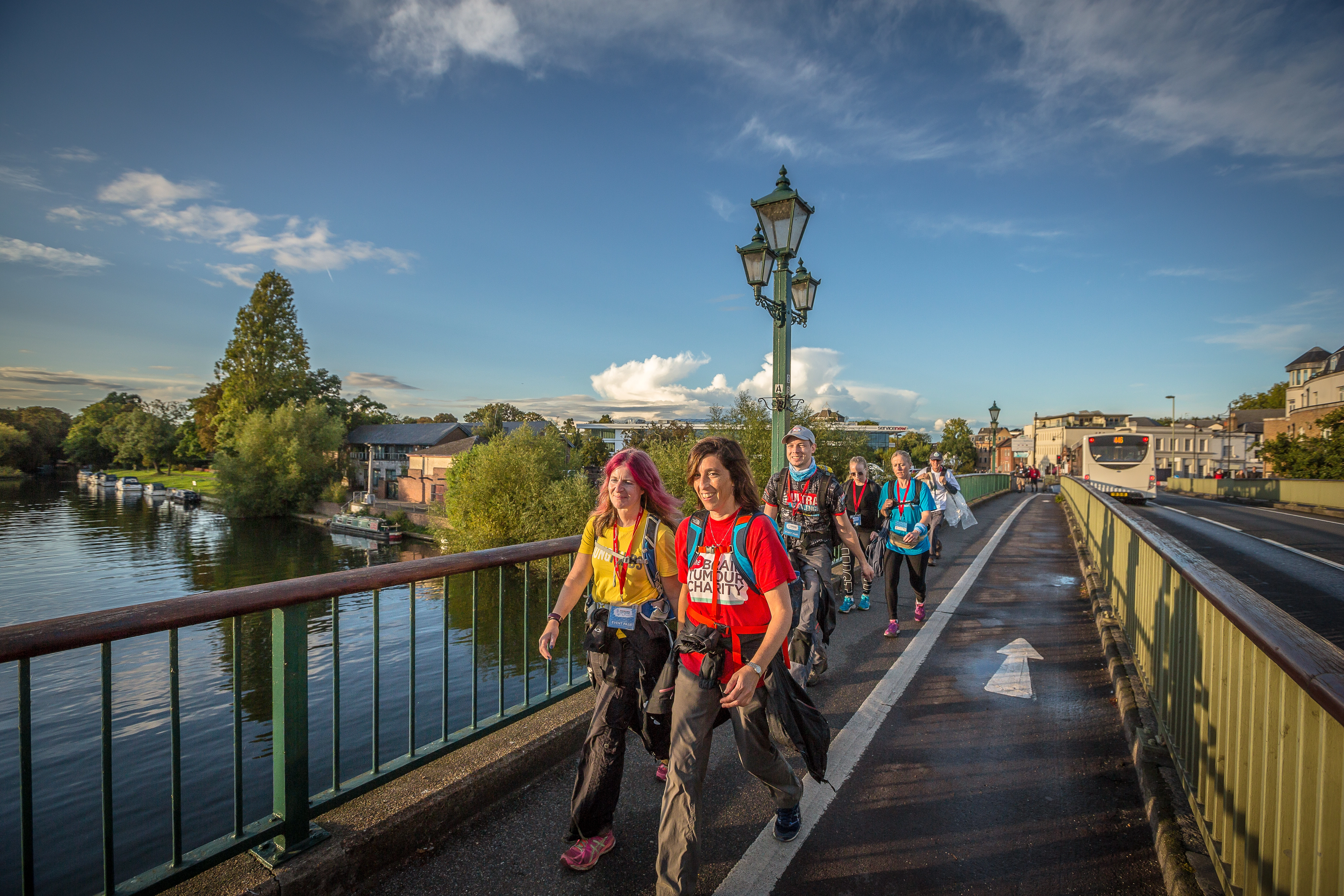 Thames Path Ultra Challenge participants walking across a bridge over a river. Participants are wearing walking gear and smiling. London's buildings and a bus can be seen the background.