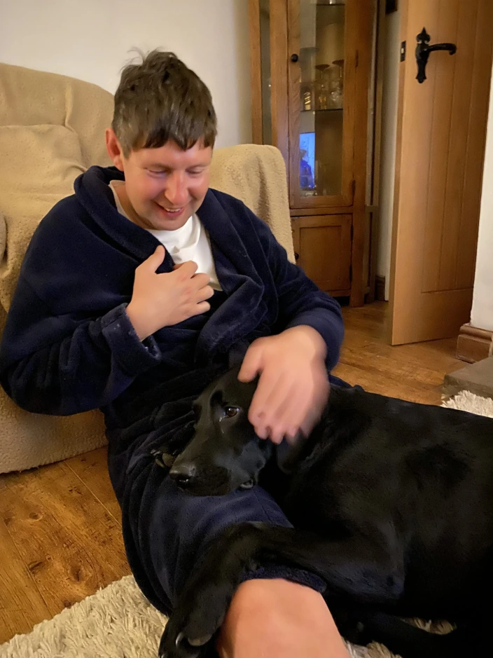 Dan sitting on the living room floor in his dressing gown smiling at black labrador Quest who is lying in his lap