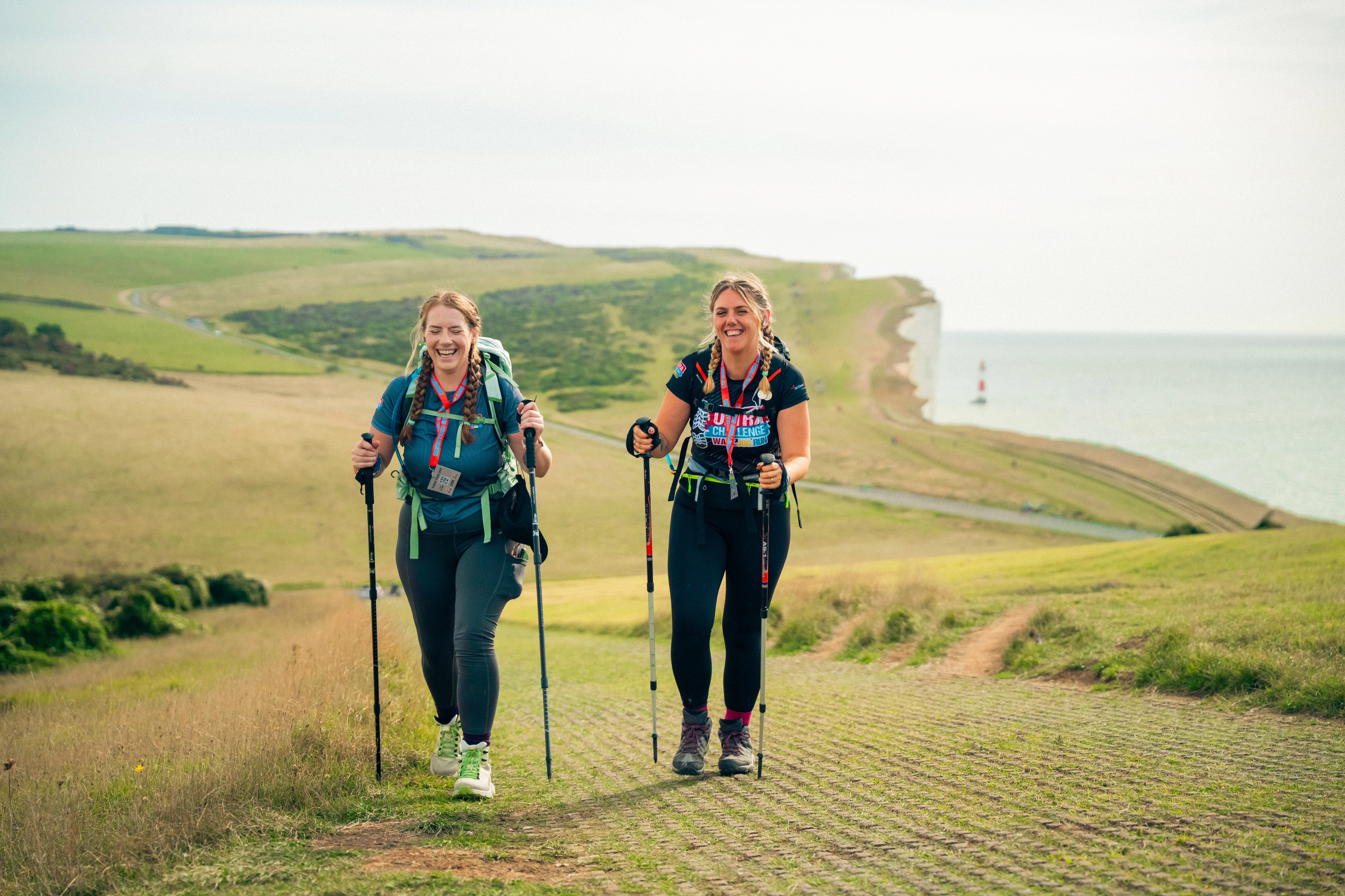 Two women walking along the coastline as part of the South Coast Ultra Challenge. They are using walking poles and smiling and laughing. Views of the sea and sea cliffs can be seen in the background.