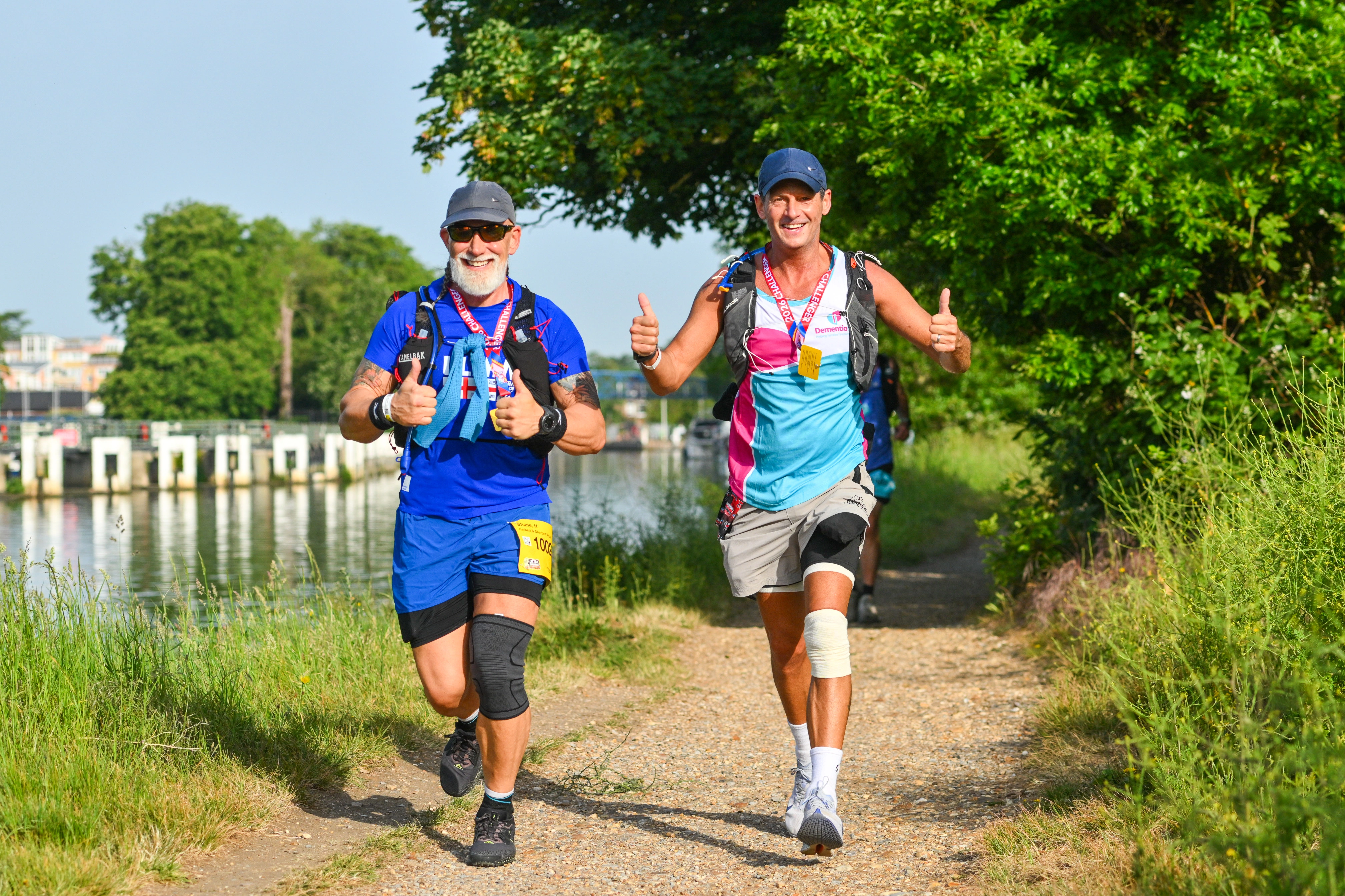 Two participants walking along a riverside nature trail as part of the London 2 Brighton Ultra Challenge. Participants are smiling and giving a thumbs up.