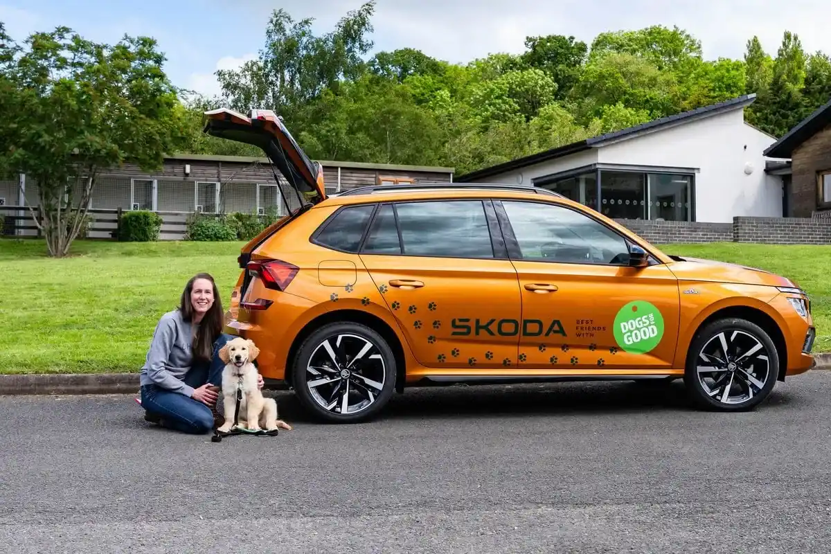 Golden retriever puppy sitting on ground next to orange Skoda car with a volunteer who is sitting next to the puppy. Grass, trees and the Dogs for Good training centre can be seen in the background