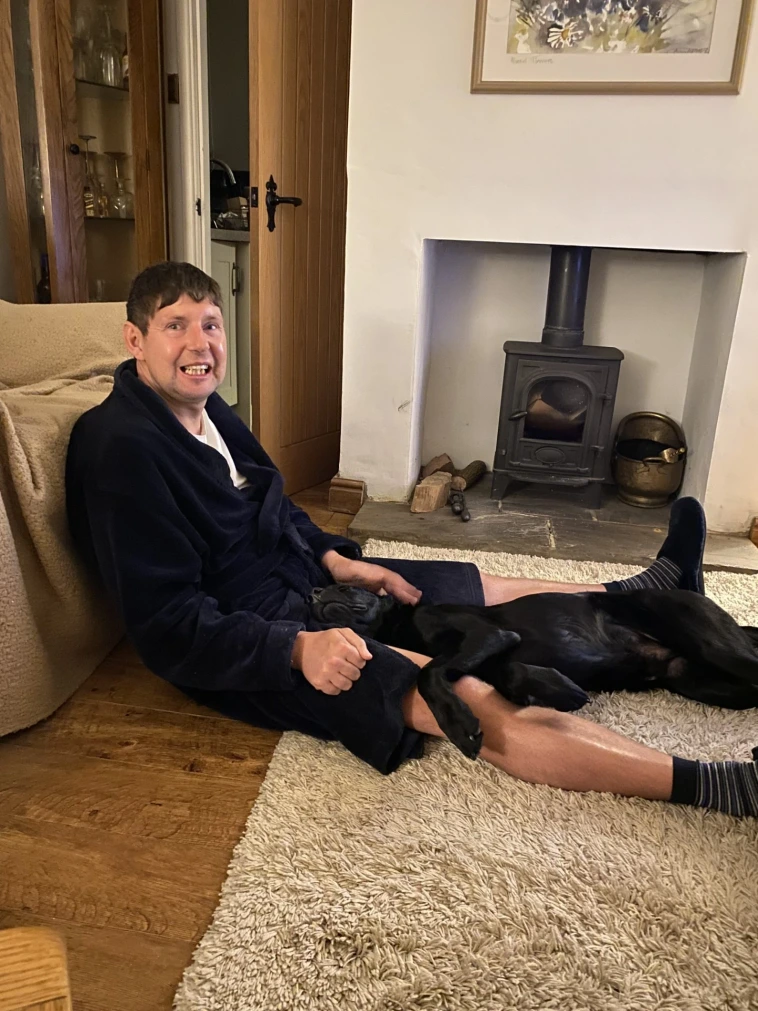 Dan and black labrador Quest on the living room floor. Dan is smiling and Quest is lying in his lap