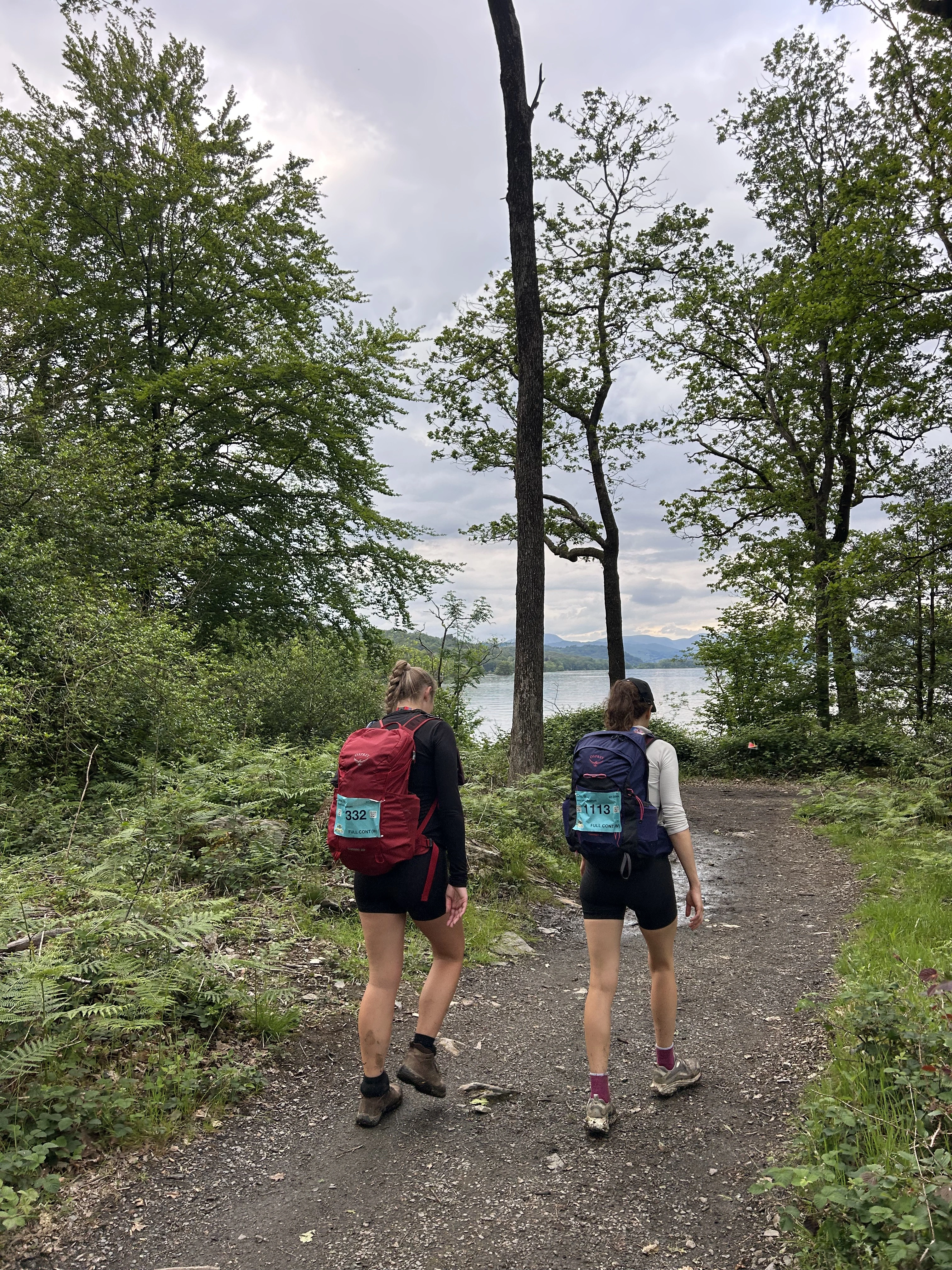 Two participants walking along a nature trail as part of the Lake District Ultra Challenge. They are wearing hiking gear and walking towards Lake Windermere, which can be seen in the background through tall trees.