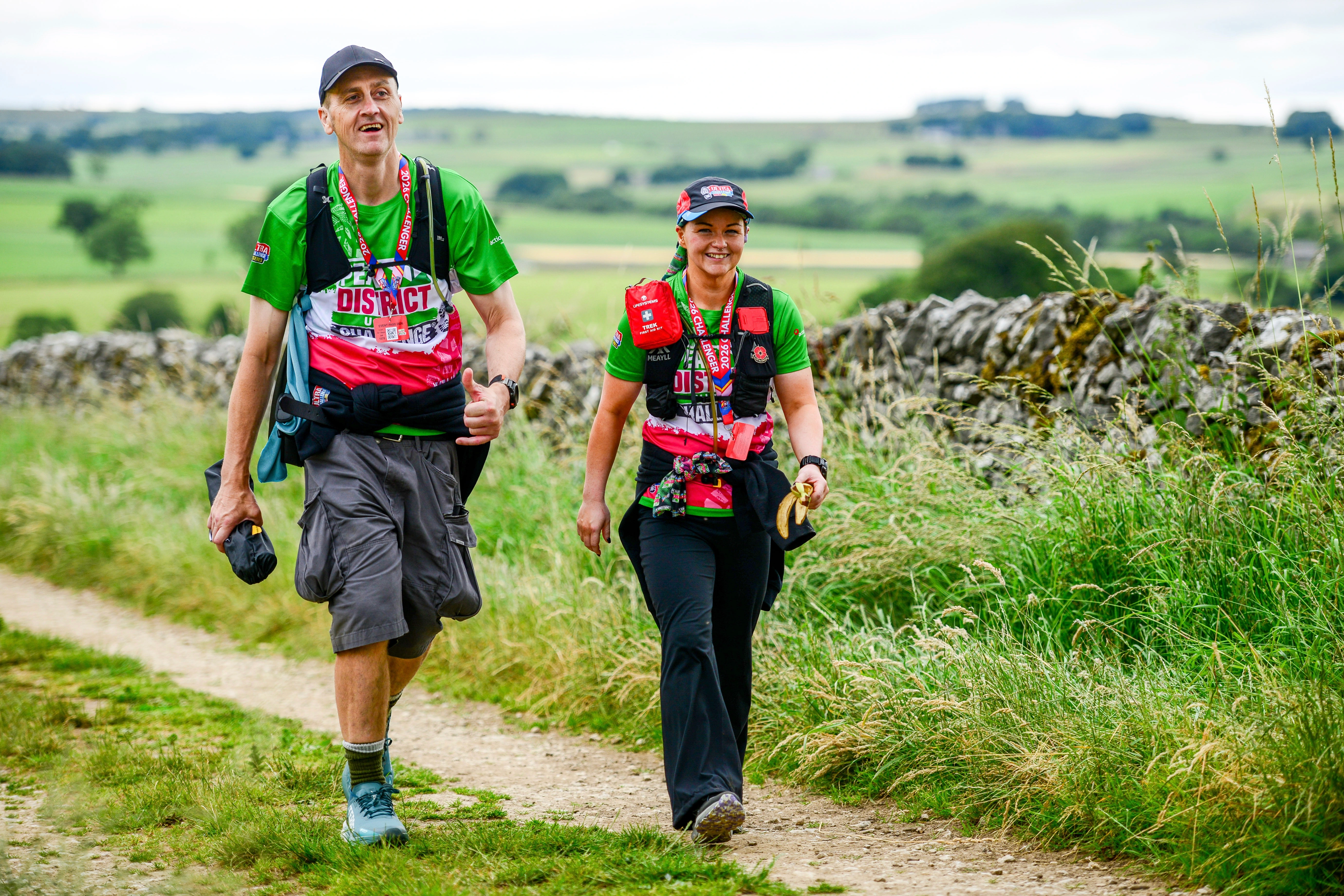 Two people walking through the Peak District as part of the Peak District Ultra Challenge. They are walking past a dry stone wall along a marked trail, trees, hills and fields can be seen in the background.