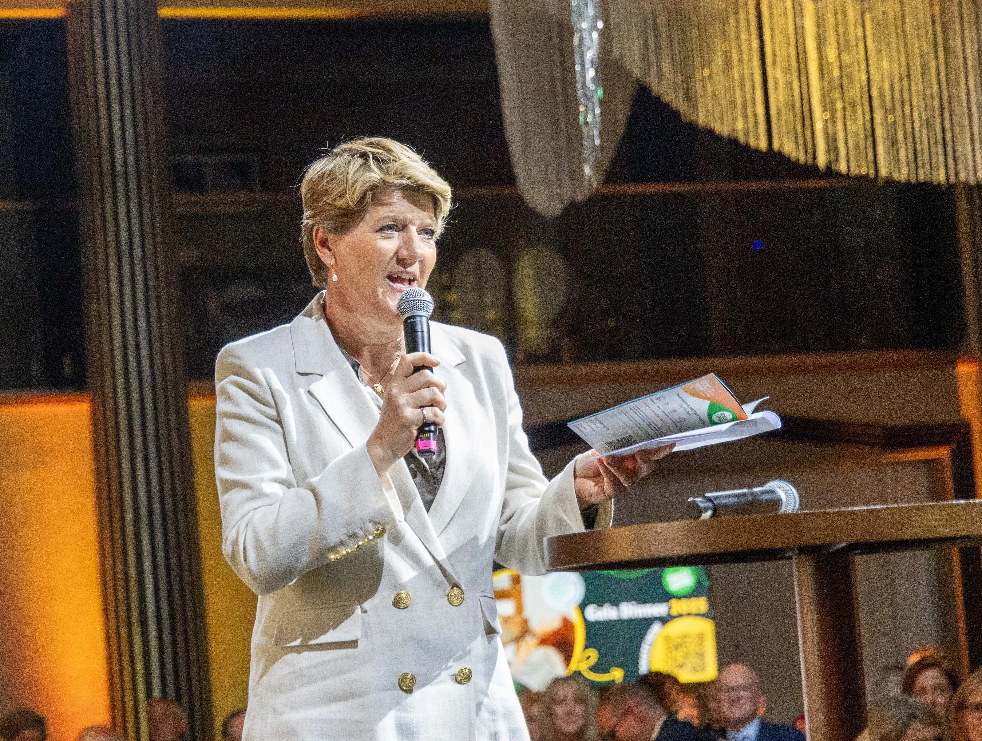 Clare Balding on stage at Dogs for Good Gala Dinner event. Clare is wearing a white suit and talking into a microphone. Crowd of attendees can be seen in the background.