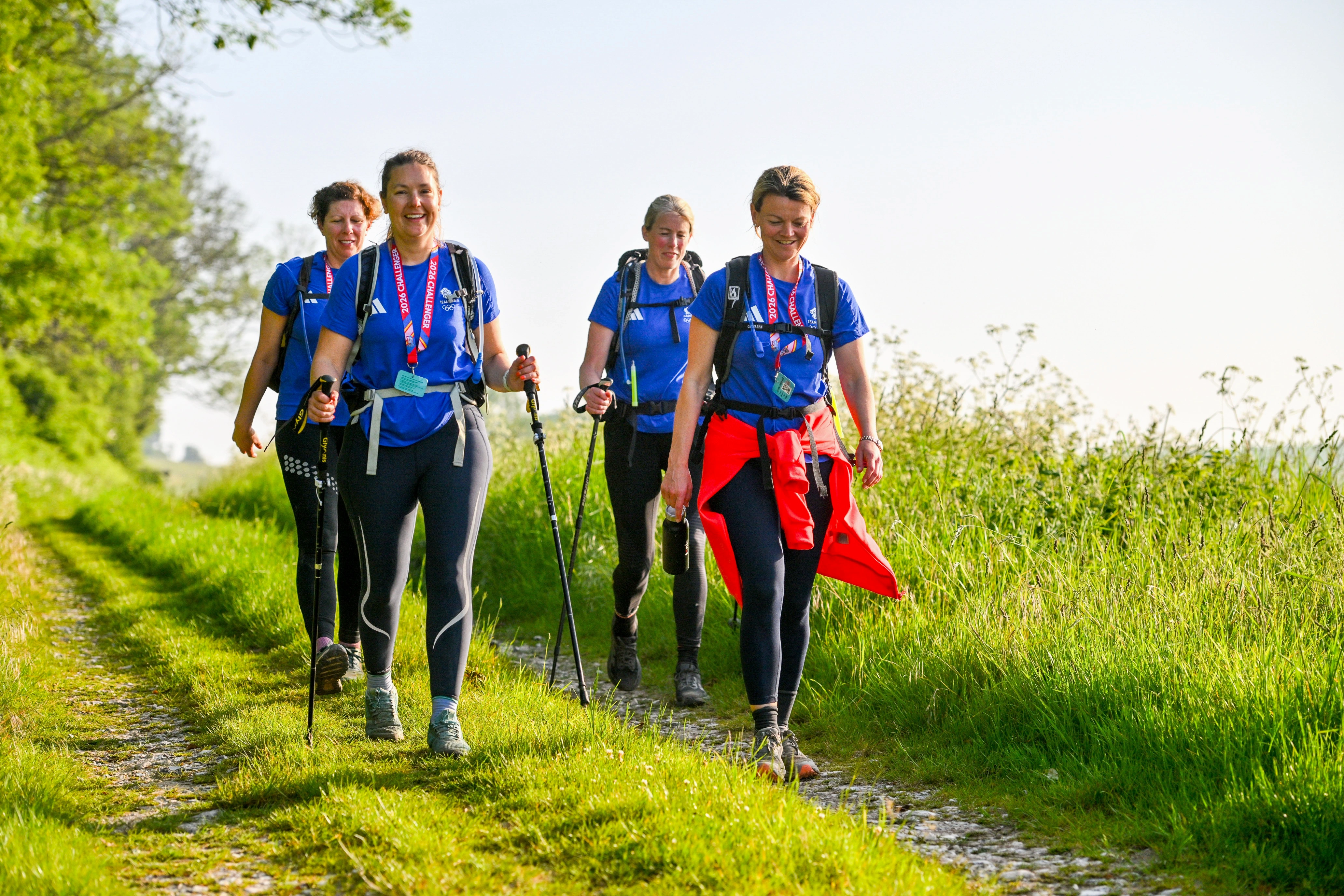 Four people in hiking clothes walking along a footpath in the sunny countryside. They are using walking poles, wearing rucksacks and smiling.