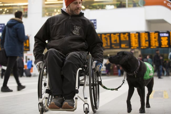 Man in wheelchair with black labrador walking alongside at train station 