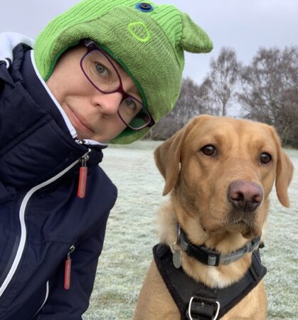 Alex is wearing a black coat zipped up, glasses and a green hat. She is stood in a field next to a light brown dog with dark brown eyes and floppy ears.