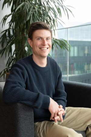 Ben is sat on a chair in front of a large leafy green plant, next to the window. He has short dark hair and is smiling. He is wearing a dark-coloured jumper and light brown trousers. 