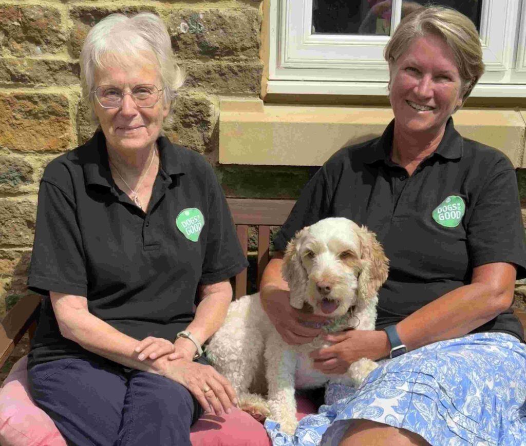 Dogs for Good volunteers Christine and Carol sit together on a sunny bench.  A cockerpoo is sat between them. 
